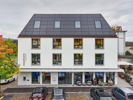 Das Bild zeigt ein dreigeschossiges Geschäftsgebäude mit einem markanten Steildach. Die Fassade ist in einem hellen Farbton gehalten und wird durch regelmässig angeordnete Fenster gegliedert, die dem Gebäude eine klare und ausgewogene Struktur verleihen. Die dunklen Fensterrahmen setzen dabei einen deutlichen Kontrast zur hellen Gebäudehülle. Das Dach ist mit dunkel ausgeführten Dachflächen und mehreren Dachfenstern gestaltet. Durch seine steile Form prägt es das Erscheinungsbild des Gebäudes und orientiert sich an der typischen Dachlandschaft des umliegenden Ortsbildes. Im Erdgeschoss sorgen grossflächige Verglasungen für eine offene und transparente Wirkung. Die Schaufensterfronten erstrecken sich über einen grossen Teil der Gebäudebreite und schaffen eine direkte Verbindung zum Strassenraum. Über den Eingangsbereichen sind Beschriftungen und Signalelemente angebracht, die auf die gewerbliche Nutzung des Gebäudes hinweisen. Das Bild zeigt ein dreigeschossiges Geschäftsgebäude mit einem markanten Steildach. Die Fassade ist in einem hellen Farbton gehalten und wird durch regelmässig angeordnete Fenster gegliedert, die dem Gebäude eine klare und ausgewogene Struktur verleihen. Die dunklen Fensterrahmen setzen dabei einen deutlichen Kontrast zur hellen Gebäudehülle. Das Dach ist mit dunkel ausgeführten Dachflächen und mehreren Dachfenstern gestaltet. Durch seine steile Form prägt es das Erscheinungsbild des Gebäudes und orientiert sich an der typischen Dachlandschaft des umliegenden Ortsbildes. Im Erdgeschoss sorgen grossflächige Verglasungen für eine offene und transparente Wirkung. Die Schaufensterfronten erstrecken sich über einen grossen Teil der Gebäudebreite und schaffen eine direkte Verbindung zum Strassenraum. Über den Eingangsbereichen sind Beschriftungen und Signalelemente angebracht, die auf die gewerbliche Nutzung des Gebäudes hinweisen.