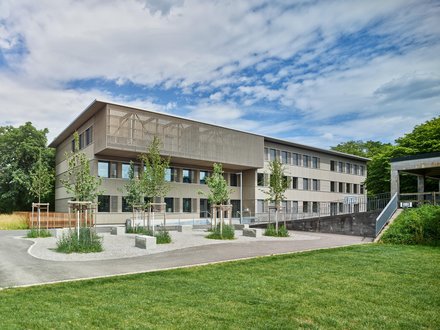Das Bild zeigt ein modernes, zweigeschossiges Gebäude in Holzmodulbauweise mit einer auffälligen Holzfassade im Obergeschoss. Vor dem Gebäude befindet sich eine Grünfläche, während das Erdgeschoss durch eine offene Gestaltung mit grossen Fensterflächen geprägt ist. Das Bild zeigt ein modernes, zweigeschossiges Gebäude in Holzmodulbauweise mit einer auffälligen Holzfassade im Obergeschoss. Vor dem Gebäude befindet sich eine Grünfläche, während das Erdgeschoss durch eine offene Gestaltung mit grossen Fensterflächen geprägt ist.