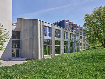 Das Bild zeigt ein modernes, mehrgeschossiges Schulgebäude, das vermutlich als Berufswahlschule oder Bildungszentrum genutzt wird. Die Architektur wirkt zeitgemäss und funktional und verbindet moderne Baumaterialien mit einer klaren, geradlinigen Gestaltung. Das Gebäude erstreckt sich über mehrere Stockwerke und besitzt eine langgezogene rechteckige Form. Die Fassade besteht überwiegend aus vertikalen Metall- oder Fassadenelementen in verschiedenen Grau- und Silbertönen. Dazwischen sind zahlreiche grosse Fensterflächen angeordnet, die für eine helle und freundliche Lernumgebung sorgen und viel Tageslicht in die Innenräume lassen. Die regelmässige Anordnung der Fenster verleiht dem Gebäude ein geordnetes und harmonisches Erscheinungsbild. Im linken Bereich des Gebäudes befindet sich ein markanter Gebäudeteil mit einer etwas geschlosseneren Fassadengestaltung. Die vertikalen Fassadenlamellen sorgen nicht nur für eine moderne Optik, sondern können auch als Sonnen- oder Sichtschutz dienen. Das Gebäude wirkt insgesamt hochwertig und nachhaltig geplant. Vor dem Schulgebäude erstreckt sich eine grosszügige Grünfläche mit dichtem, gepflegtem Bewuchs. Die Wiese oder bepflanzte Fläche bedeckt einen grossen Teil des Vordergrunds und schafft einen angenehmen Kontrast zur modernen Architektur. Auf der rechten Seite steht ein grosser Laubbaum mit dichter Krone, der das Schulareal zusätzlich aufwertet und für eine naturnahe Atmosphäre sorgt. Die Aufnahme entstand bei sonnigem Wetter. Der blaue Himmel und die helle Beleuchtung lassen das Gebäude freundlich und einladend erscheinen. Die Sonnenstrahlen erzeugen leichte Schatten auf der Fassade und betonen die Struktur der vertikalen Fassadenelemente. Insgesamt vermittelt das Gebäude den Eindruck einer modernen Bildungsstätte, die sowohl funktionale Anforderungen als auch architektonische Qualität berücksichtigt. Die Kombination aus klarer Architektur, grosszügigen Fensterflächen und den umgebenden Grünanlagen schafft ein angenehmes Umfeld für Lernen, Ausbildung und persönliche Entwicklung. Das Bild zeigt ein modernes, mehrgeschossiges Schulgebäude, das vermutlich als Berufswahlschule oder Bildungszentrum genutzt wird. Die Architektur wirkt zeitgemäss und funktional und verbindet moderne Baumaterialien mit einer klaren, geradlinigen Gestaltung. Das Gebäude erstreckt sich über mehrere Stockwerke und besitzt eine langgezogene rechteckige Form. Die Fassade besteht überwiegend aus vertikalen Metall- oder Fassadenelementen in verschiedenen Grau- und Silbertönen. Dazwischen sind zahlreiche grosse Fensterflächen angeordnet, die für eine helle und freundliche Lernumgebung sorgen und viel Tageslicht in die Innenräume lassen. Die regelmässige Anordnung der Fenster verleiht dem Gebäude ein geordnetes und harmonisches Erscheinungsbild. Im linken Bereich des Gebäudes befindet sich ein markanter Gebäudeteil mit einer etwas geschlosseneren Fassadengestaltung. Die vertikalen Fassadenlamellen sorgen nicht nur für eine moderne Optik, sondern können auch als Sonnen- oder Sichtschutz dienen. Das Gebäude wirkt insgesamt hochwertig und nachhaltig geplant. Vor dem Schulgebäude erstreckt sich eine grosszügige Grünfläche mit dichtem, gepflegtem Bewuchs. Die Wiese oder bepflanzte Fläche bedeckt einen grossen Teil des Vordergrunds und schafft einen angenehmen Kontrast zur modernen Architektur. Auf der rechten Seite steht ein grosser Laubbaum mit dichter Krone, der das Schulareal zusätzlich aufwertet und für eine naturnahe Atmosphäre sorgt. Die Aufnahme entstand bei sonnigem Wetter. Der blaue Himmel und die helle Beleuchtung lassen das Gebäude freundlich und einladend erscheinen. Die Sonnenstrahlen erzeugen leichte Schatten auf der Fassade und betonen die Struktur der vertikalen Fassadenelemente. Insgesamt vermittelt das Gebäude den Eindruck einer modernen Bildungsstätte, die sowohl funktionale Anforderungen als auch architektonische Qualität berücksichtigt. Die Kombination aus klarer Architektur, grosszügigen Fensterflächen und den umgebenden Grünanlagen schafft ein angenehmes Umfeld für Lernen, Ausbildung und persönliche Entwicklung.