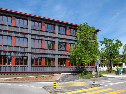 Das moderne Schulgebäude überzeugt durch seine klare, zeitgemässe Architektur und die grosszügig gestalteten Fensterflächen, die für helle und freundliche Lernräume sorgen. Die markante Fassadengestaltung mit roten Akzenten verleiht dem Gebäude einen eigenständigen Charakter und setzt architektonische Akzente. Die transparente Gebäudehülle schafft einen hohen Tageslichteinfall und fördert eine angenehme Lernatmosphäre. Die umgebenden Grünflächen und die sorgfältig gestaltete Aussenanlage integrieren das Schulgebäude harmonisch in sein Umfeld. Das moderne Schulgebäude überzeugt durch seine klare, zeitgemässe Architektur und die grosszügig gestalteten Fensterflächen, die für helle und freundliche Lernräume sorgen. Die markante Fassadengestaltung mit roten Akzenten verleiht dem Gebäude einen eigenständigen Charakter und setzt architektonische Akzente. Die transparente Gebäudehülle schafft einen hohen Tageslichteinfall und fördert eine angenehme Lernatmosphäre. Die umgebenden Grünflächen und die sorgfältig gestaltete Aussenanlage integrieren das Schulgebäude harmonisch in sein Umfeld.