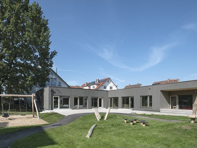 Kindertagesstätte, Fischbach - Rückansicht des Gebäudes mit grauer Fassade und großer Außenanlage mit Spielgeräten aus Holz 