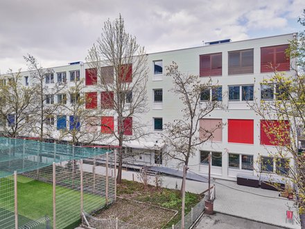 Das Bild zeigt ein modernes, mehrstöckiges Gebäude mit einer hellen Fassade in Weiss- und Grautönen. Auffällig sind die roten Fensterrahmen und Fassadenelemente, die dem Gebäude ein lebendiges Erscheinungsbild verleihen. Die Architektur ist geradlinig und funktional, mit grossen Fensterflächen, die auf eine Nutzung als Schule, Kindergarten oder öffentliches Gebäude hindeuten.  Im Vordergrund befindet sich eine grüne Rasenfläche. Davor verläuft ein Metallzaun, der das Grundstück begrenzt. Mehrere Bäume mit dichtem Laub stehen zwischen Kamera und Gebäude. Die Äste und Blätter verdecken Teile der Fassade und verleihen dem Bild eine natürliche Rahmung.  Das Gebäude erstreckt sich über eine grössere Länge und besitzt mindestens drei Geschosse. Das Dach ist flach gestaltet und weist ebenfalls rote Akzente auf. Die Aufnahme wurde bei Tageslicht gemacht; die Beleuchtung wirkt gleichmässig und die Farben erscheinen natürlich.