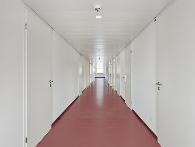 Limmatprovisorium, Zürich - Langer schmaler Korridor eines Holz-Modulbaus mit rotem Boden und geschlossenen Türen zu Büroräumlichkeiten.