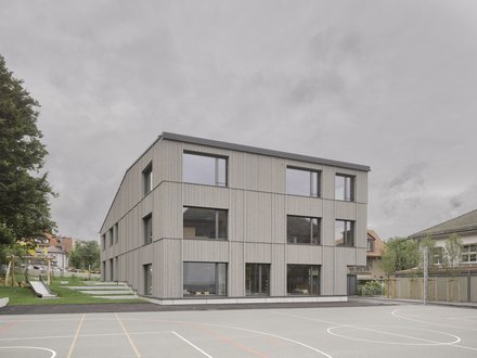 Zweigeschossiges Schulgebäude in Hybridbauweise mit grauer Fassade und grossen Fenstern.