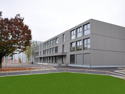 Schulgebäude Horburg, Basel - Zweigeschossiges Schulgebäude mit grauer Fassade und einer grossen grüner Wiese.