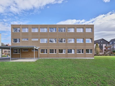 Modernes Schulhaus in Holzbauweise mit klarer, zeitgemässer Architektur. Das mehrgeschossige Gebäude überzeugt durch grosse Fensterflächen, die helle und freundliche Lernräume ermöglichen. Die horizontale Fassadengestaltung aus Holz verleiht dem Schulhaus eine natürliche und nachhaltige Ausstrahlung, während die grosszügigen Aussenflächen vielfältige Nutzungsmöglichkeiten für Schule und Freizeit bieten.