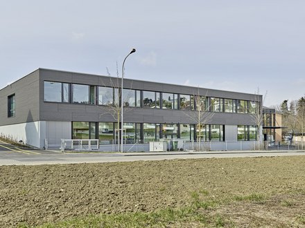 Heilpädagogische Schule Bülach, Winkel - Zweigeschossiges Schulgebäude mit grauer Holzfassade und grosszügigen Fenster.