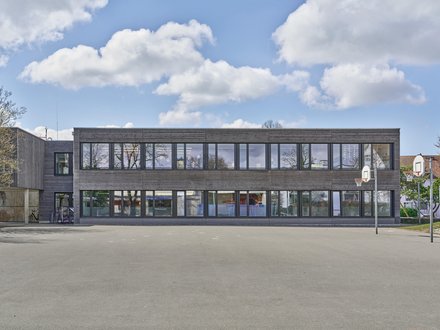 Das Bild zeigt ein modernes, zweigeschossiges Schulgebäude in zeitgemässer Architektur. Der langgestreckte Baukörper verfügt über eine klare, horizontale Gestaltung mit einer Kombination aus grossflächigen Verglasungen und dunklen Fassadenelementen. Die Konstruktion wirkt leicht und offen und lässt auf eine Holzbauweise schliessen. Im Erdgeschoss befinden sich grosszügige Fensterfronten, die viel Tageslicht in die Unterrichtsräume bringen. Das Obergeschoss zeichnet sich durch ein regelmässiges Fassadenraster mit breiten Fensterbändern aus. Die Fassade präsentiert sich in einem modernen, zurückhaltenden Farbkonzept mit dunklen und natürlichen Materialien. Vor dem Gebäude liegt ein grosszügiger Pausen- oder Vorplatzbereich mit befestigter Oberfläche. Im Hintergrund sind weitere Gebäudeteile sowie einzelne Bäume erkennbar. Der Himmel ist teilweise bewölkt, wodurch das Gebäude in einem hellen und freundlichen Licht erscheint. Das Bild zeigt ein modernes, zweigeschossiges Schulgebäude in zeitgemässer Architektur. Der langgestreckte Baukörper verfügt über eine klare, horizontale Gestaltung mit einer Kombination aus grossflächigen Verglasungen und dunklen Fassadenelementen. Die Konstruktion wirkt leicht und offen und lässt auf eine Holzbauweise schliessen. Im Erdgeschoss befinden sich grosszügige Fensterfronten, die viel Tageslicht in die Unterrichtsräume bringen. Das Obergeschoss zeichnet sich durch ein regelmässiges Fassadenraster mit breiten Fensterbändern aus. Die Fassade präsentiert sich in einem modernen, zurückhaltenden Farbkonzept mit dunklen und natürlichen Materialien. Vor dem Gebäude liegt ein grosszügiger Pausen- oder Vorplatzbereich mit befestigter Oberfläche. Im Hintergrund sind weitere Gebäudeteile sowie einzelne Bäume erkennbar. Der Himmel ist teilweise bewölkt, wodurch das Gebäude in einem hellen und freundlichen Licht erscheint.