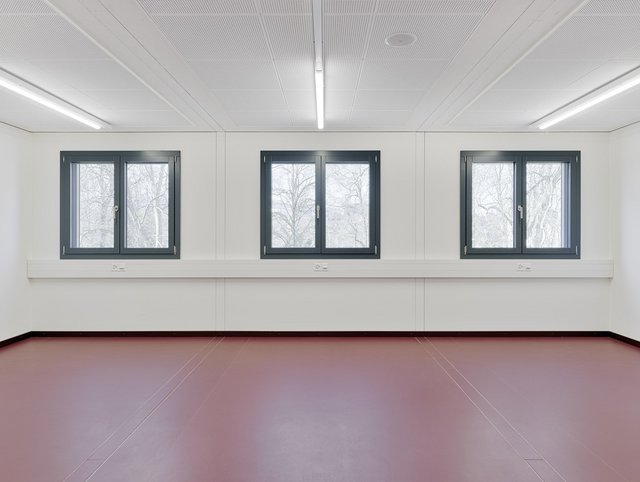 Limmatprovisorium, Zürich - Leerer Raum eines Modulbau Provisoriums mit rotem Boden und drei Fenstern mit dunklem Rahmen.