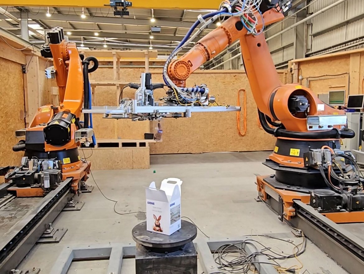 Zwei organge Fassadenroboter in einer Fertigungshalle. Der eine Roboter befüllt eine kleine weisse Osterkiste mit einem Schokoladenhase.