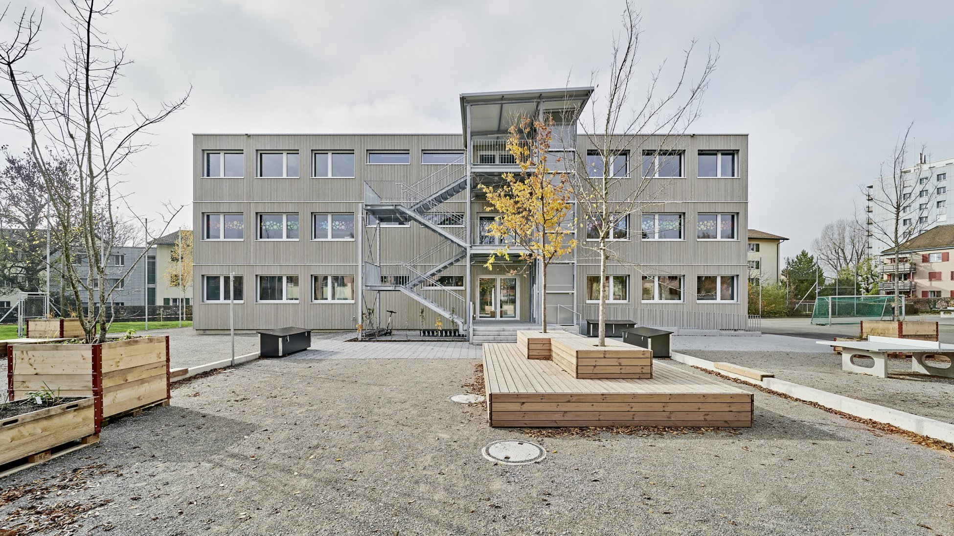 3-geschossiger Modulbau mit hellgrauer Holzfassade und grosszügigem Pausenplatz mit Sitzmöglichkeiten in Holz