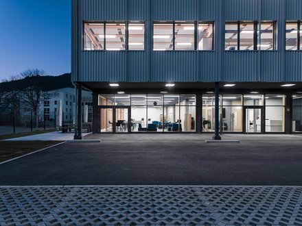 Brütsch Elektronik, Beringen  - Aussenansicht des Eingangsbereichs bei Nacht, mit hell erleuchteten Fenster; Stahlstützen und Fassade erkennbar. 