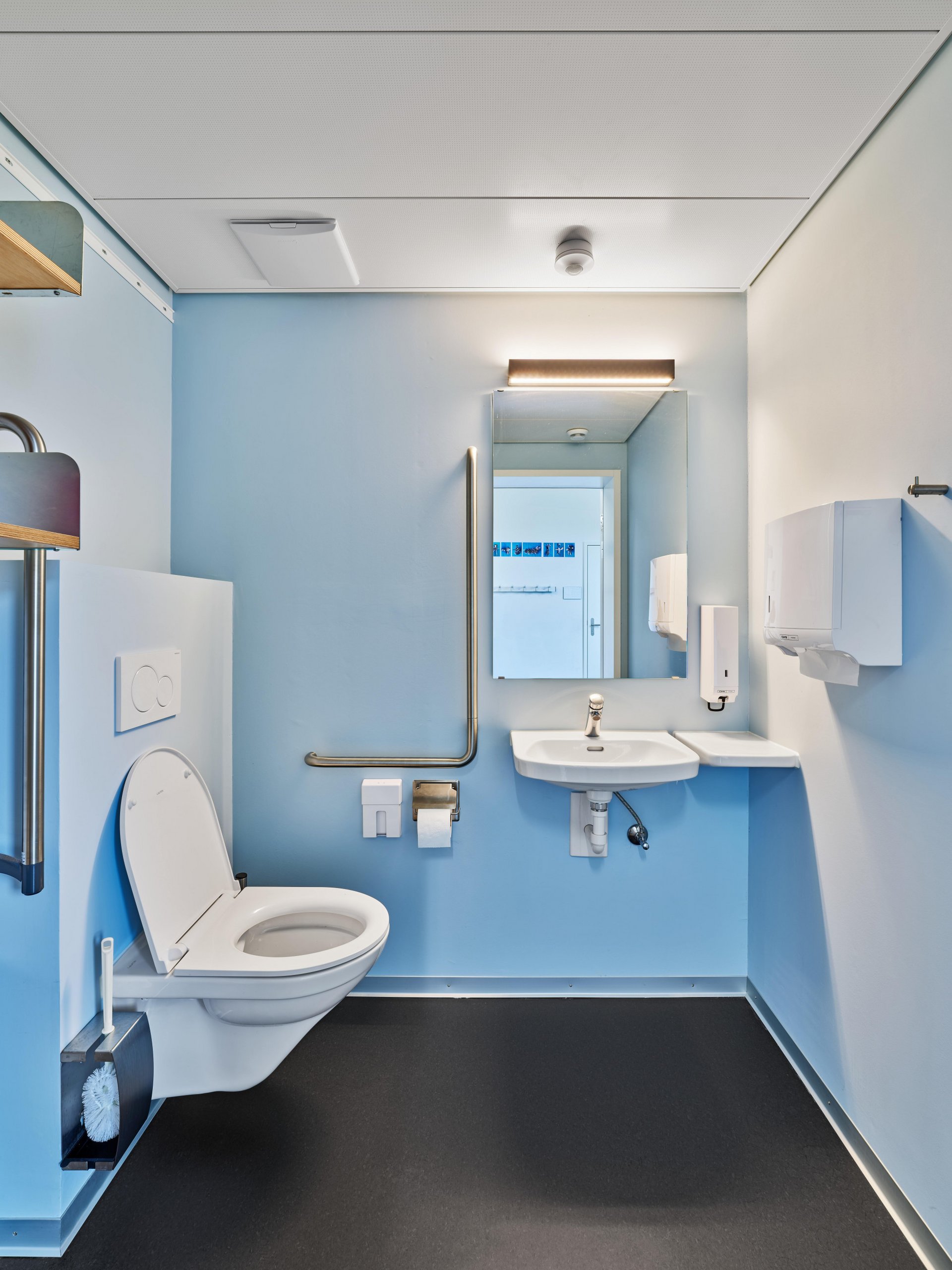 Das Bild zeigt einen modernen und barrierearmen Sanitärraum mit hellen Farben und einer funktionalen Ausstattung.

Im Vordergrund links befindet sich ein wandmontiertes WC mit Haltegriffen auf beiden Seiten. Die Haltegriffe erleichtern die Nutzung und deuten darauf hin, dass es sich um eine behindertengerechte Toilette handelt. Neben dem WC ist ein Toilettenpapierhalter angebracht.

Im hinteren Bereich des Raumes befindet sich ein Waschbecken mit darunter freiem Beinraum, sodass es auch mit einem Rollstuhl gut zugänglich ist. Über dem Waschbecken hängt ein grosser Spiegel. Rechts daneben sind ein Seifenspender und ein Papierhandtuchspender an der Wand montiert.

Die Wände sind teilweise weiss und teilweise in einem hellen Blauton gehalten. Auch die Tür zum Sanitärbereich ist hellblau. Der Boden ist dunkel und bildet einen deutlichen Kontrast zu den hellen Wandflächen. Die Decke besteht aus weissen Akustikplatten mit integrierter Beleuchtung.

Im Spiegel sind weitere Teile des Raumes sowie eine zusätzliche Tür sichtbar. Insgesamt wirkt der Sanitärraum modern, sauber, hell und auf die Anforderungen der Barrierefreiheit ausgelegt.